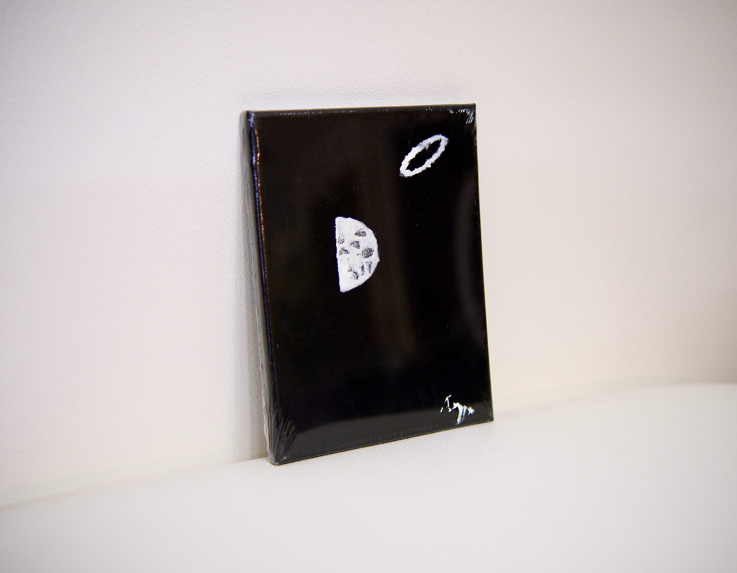 The small black canvas propped at an angle against a white wall, its glossy surface catching the light; the white half-moon and ring read faintly across it.