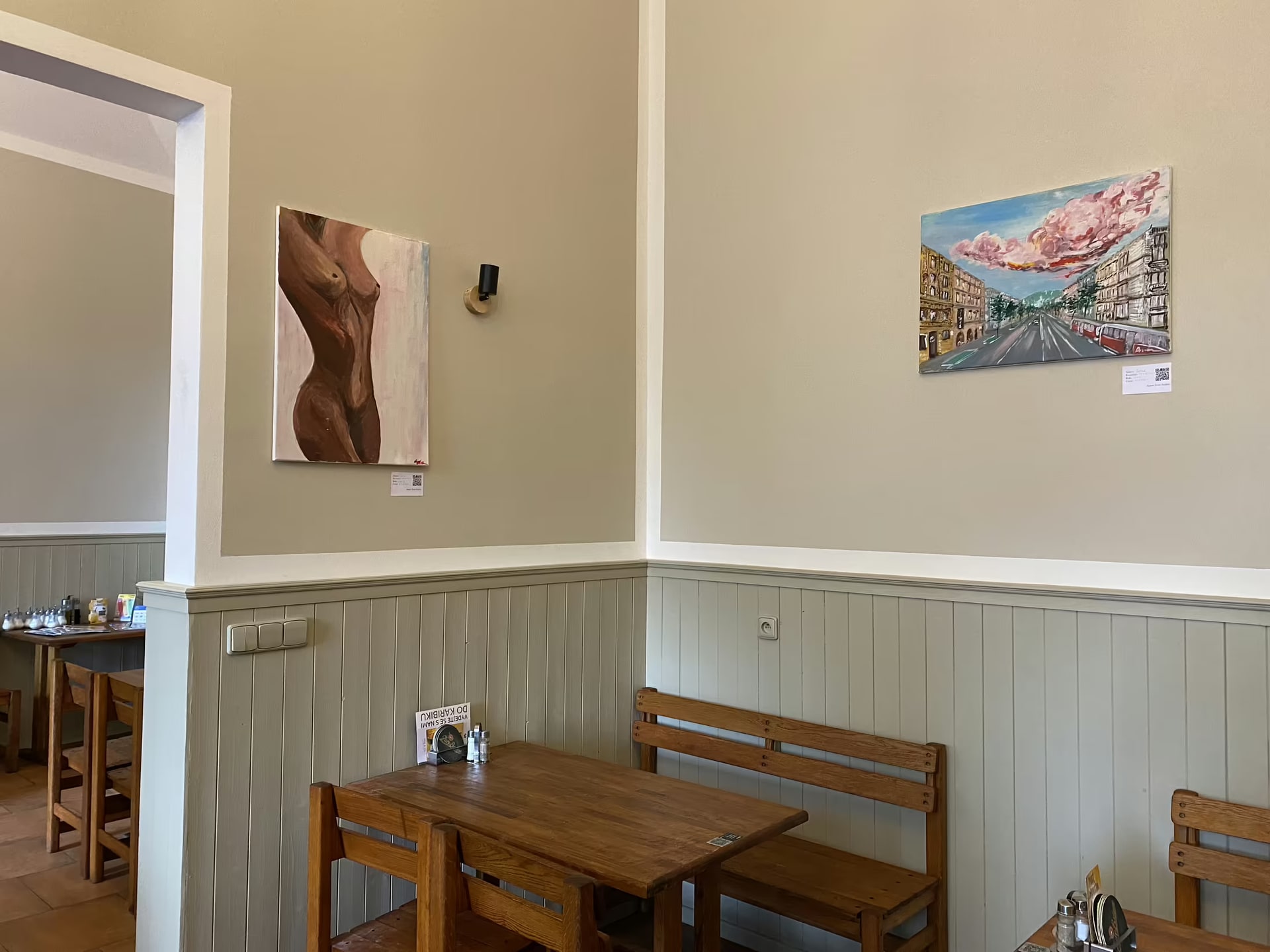 Two paintings by Ivan Anikin installed on the cream walls of a Prague cafe above wooden tables.